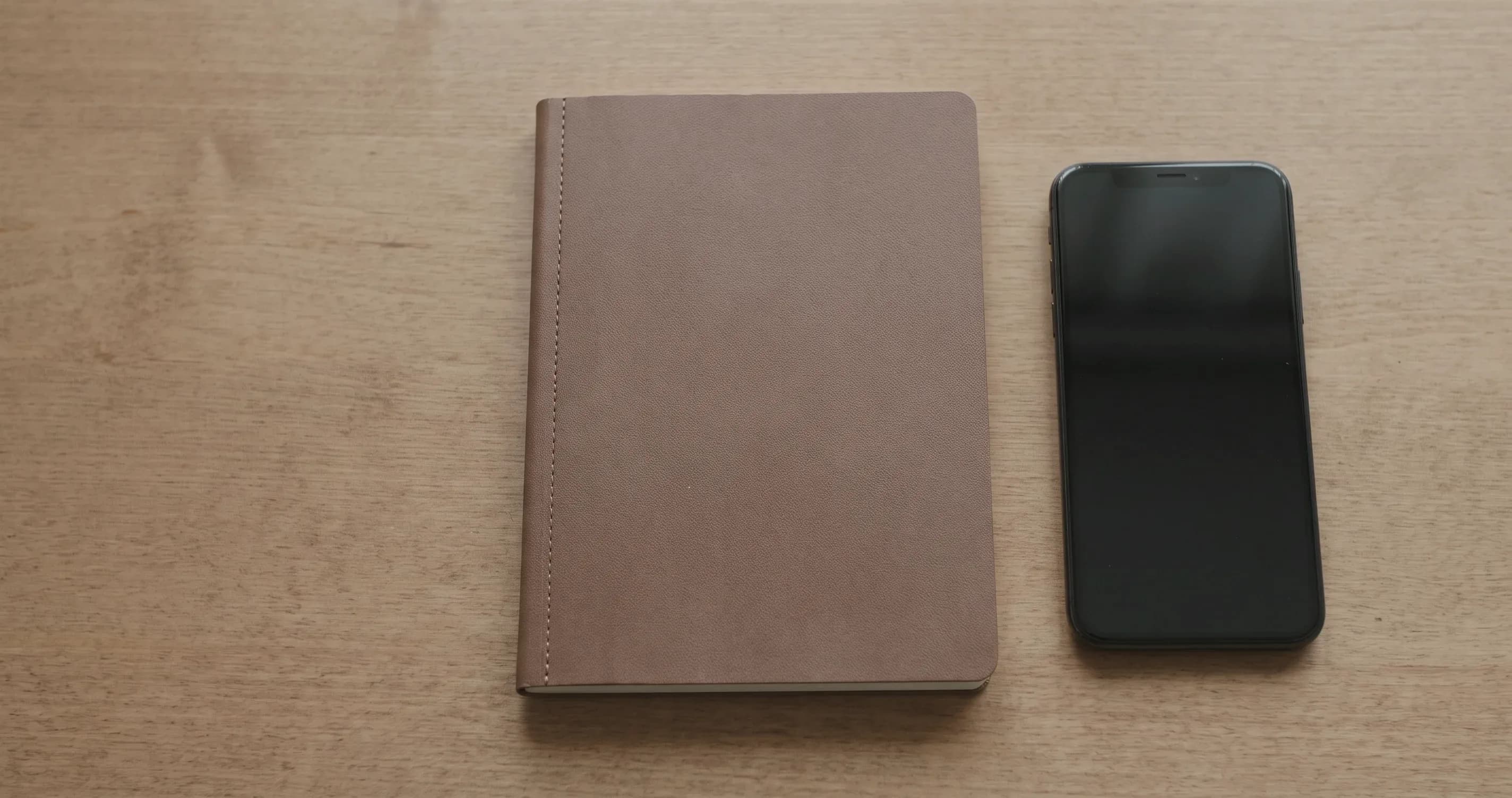 A closed notebook lying beside a switched-off phone on a wooden table.