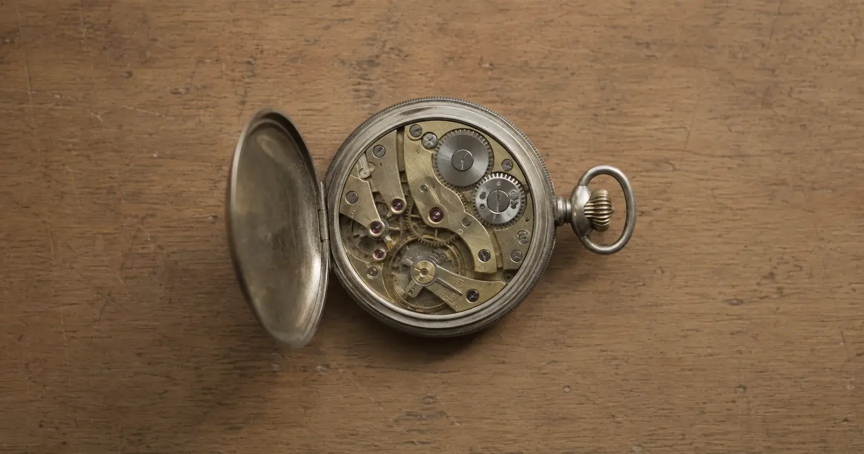 An antique pocket watch on a wooden bench, its cover hinged open to expose the movement inside.