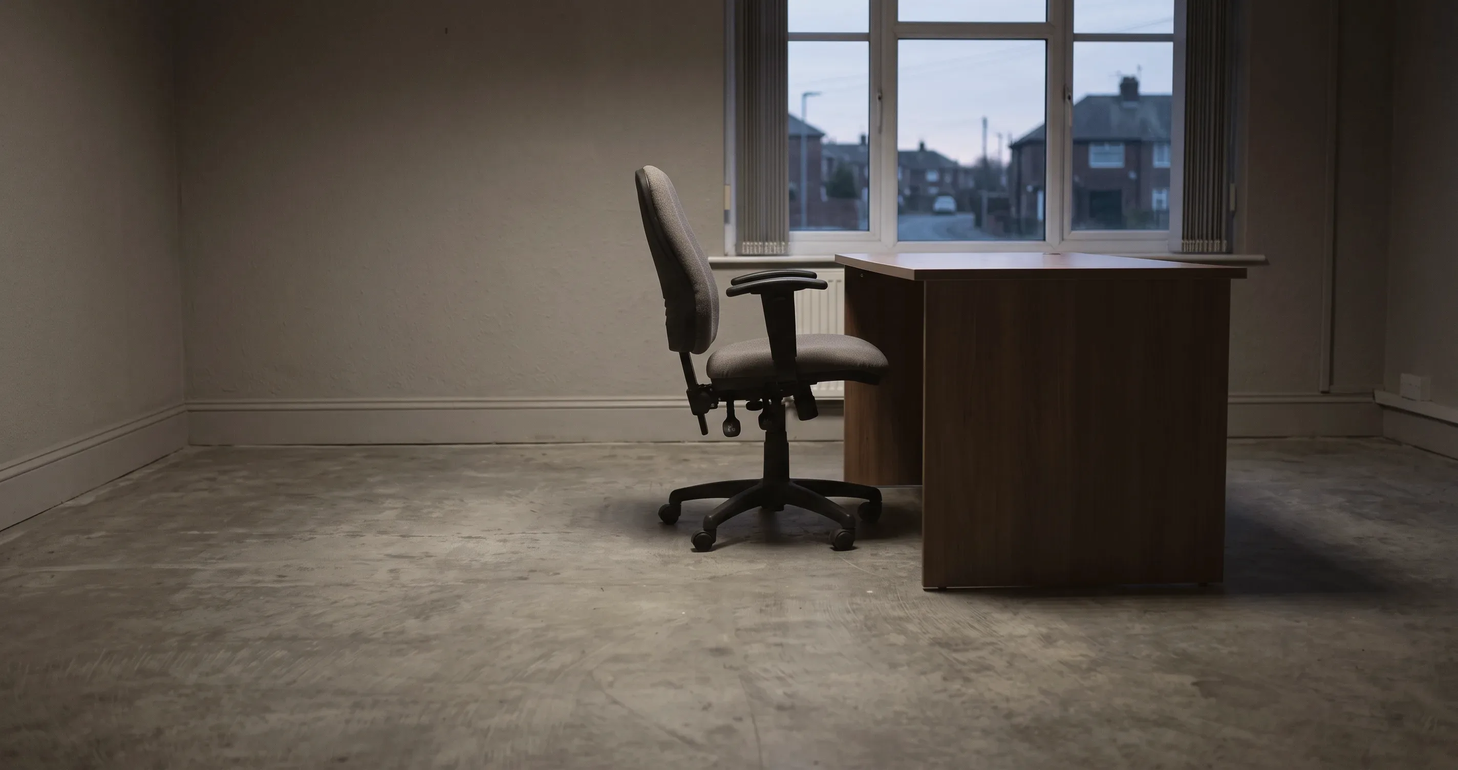 An empty desk chair pushed back from a bare desk in an office at dusk.